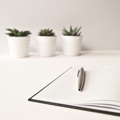 Symbolbild. Notizblock, Kugelschreiber auf einem weißen Schreibtisch. Im Hintergrund 3 kleine Kakteen