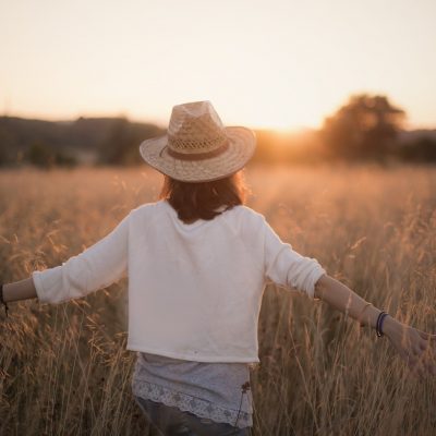 Titelbild zu Vitalissimo. Sonnenuntergang, Frau läuft mit ausgebreiteten Armen durch ein Feld. Sie trägt Jeans, eine weiße Bluse und einen Hut. Im Hintergrund die untergehende Sonne und ein Baum.